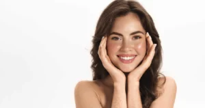 Smiling young woman with clear, glowing skin and natural makeup, gently touching her face with both hands against a white background in Kentfield, CA, after Radiesse Treatments.