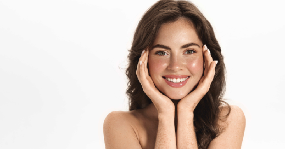 Smiling young woman with clear, glowing skin and natural makeup, gently touching her face with both hands against a white background in Kentfield, CA, after Radiesse Treatments.